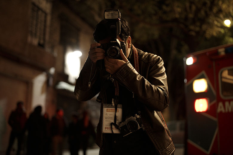 Habiendo dirigido dos exitosas comedias, ambas incorporadas al catálogo de Netflix, Luis Javier Henaine da un giro completamente distinto y se adentra de lleno al cine de terror con Desaparecer por completo, su tercer largometraje y el segundo que estrena dentro de la sección de largometraje mexicano del Festival Internacional de Cine de Morelia. Una película que transforma las agresiones visuales y la tortura mediática de la violencia en México en costras físicas en el cuerpo del avaro insensible que capitaliza de ellas.  En sus dos largometrajes previos Solteras y Tiempos felices, ambosprotagonizados por Cassandra Ciangherotti y ambos valiéndole una nominación a los premios Ariel a mejor actriz, Henaine parecía haber establecido su tema de interés al centrar, hasta ese momento, su filmografía en la industria del amor y en la obsesión (más de nuestro siglo) de controlar las relaciones y vínculos afectivos cual trámite burocrático.   En Tiempos felices era con un joven incapaz de comunicarse con su pareja para terminar la relación que recurría a una agencia interventora que, como oficina de recursos humanos que despide y amenaza en nombre de un jefe, ofrece hacer la tarea por él.   En Solteras con una mujer víctima de la presión social que, dispuesta a llegar a los 30´s casada, se adentra al intensivo curso de una casamentera en busca del manual fácil y digerible que conduzca una relación exitosa al matrimonio.  En ambas, sus protagonistas decepcionadas de los resultados de sus cursos y empresas terminan comprendiendo la complejidad de la interacción humana que no puede reducirse a una transacción económica o a un organigrama empresarial. Y digo que Henaine parecía tener ese interés porque ambas películas además de la línea temática llegaban a la misma conclusión. Ahora podría argumentarse que Desaparecer por completo trata de la industria sensacionalista de la muerte y de quienes la conforman; policías y personal de morgue corruptos, fotógrafos insensibles, periódicos amarillistas, noticieros escuetos y el consumidor morboso, sin embargo, Henaine es más incisivo y represor con quien se aprovecha de la muerte. Es muy distinto quien vende corazones inflados de helio, chocolates y la falsa ilusión de los estímulos a quien pone nuestros cuerpos en venta y arrebata nuestra dignidad.   Harold Torres interpreta a Santiago, un fotógrafo de nota roja con ambiciones de artista. En la secuencia inicial toma una fotografía de una mujer recién atropellada a la que manipula él personalmente buscando lograr la mejor composición posible de una situación estrepitosa, dejando ver que busca una apreciación estética de su obra. La fotografía que toma es un guiño a la muy famosa de EnriqueMetinides de 1979 de la periodista Adela Legarreta Rivas atropellada y prensada entre dos postes de luz en la Colonia Roma.  No sé sabe si Metinides es una inspiración de Santiago dentro de la ficción, de Henaine sí. Santiago a diferencia de Metinides no encuentra el éxito ni la gloria como fotógrafo. Su jefe incomprensivo en el periódico le reclama los largos tiempos de entrega y sus “pretensiones” que terminan cortándole; quiere que se limite a las fotografías crudas y prácticas, o cómo él mismo dice “déjate de joterias”. Para él, Santiago paga pobremente sus logros y desprecia sus halagos porque ¿qué mayor reconocimiento puede un fotógrafo alcanzar que aparecer en la primera plana?  Del otro lado las instituciones culturales y galerías de arte, aquellas que validan quien es artista y quien no, lo rechazan incesantemente. Para unos un pretencioso, para otros un vulgar sin mayor mérito.  Cuando su jefe promete financiarle su propia exposición como un incentivo para que se esmere en conseguir las imágenes más gráficas posibles, Santiago saca su instinto carroñero y aquí comienza el tema de la película de Henaine.  Santiago ha creado una rutina muy específica y cuidada para lograr capturar las más sórdidas imágenes; los policías son sus aliados quienes le informan de accidentes y le dan prioridad en la escena, conoce los lugares a los que entra por lo que no tiene reparo de saltar ventanas, subir por balcones o rodear jardines, se comunica a través de un sistema radial similar al de la policía y ha sacrificado sus días al descanso para mantenerse en las noches despierto aunque eso involucre el contacto mínimo con su pareja (quien lo resiente).  Harold Torres remite inmediatamente al Jake Gyllenhall de Nightcrawler, sin embargo, su ambición a diferencia de la de Gyllenhaal sí encuentra tope cuando tras fotografiar lo poco que queda del cuerpo carcomido por las ratas de un gobernador, una maldición, casi como castigo divino, cae sobre él haciendo que paulatinamente pierda uno a uno los sentidos. En un inicio una bendición, pues la desensibilización moral que le permite fotografiar trozos humanos como bolsas de basura se conjunta con la física y le ayuda a estar justo al lado de restos putrefactos sin el mínimo asco, pronto se convierte en la amenaza de desaparecer.  Gyllenhaal en personaje es la cara de la inhumanidad, con un cuerpo mancillado, desnutrido y débil que acompaña a un esqueleto que está por expulsar sus dos globos oculares en cualquier momento. Su físico espeluznante y la repulsión que despierta en sus allegados de gremio son las advertencias al sadismo y el morbo, que al agredir todo a nuestro paso terminamos infringiéndolo también a nosotros mismos hasta ya no ser humanos, solo bolsas de basura que tratamos con la misma consideración y dignidad que a las que grabamos en cámara.  Mismo tema y en otro contexto, Henaine habla también de la ligera línea que hay entre observador y agresor. A él no le interesa la transformación psicológica de su personaje como en Nightcrawler sino en la física del castigo que un brujo le ha impuesto. Como en Huesera, de Michelle Garza Cervera, Desaparecer por completo está inserta en su contexto, con todas las pautas y convencionalismos bien establecidos por el cine de terror gringo más comercial, pero caminando en pies muy claros sobre el folclor mexicano que quiere abordar.   La sola mención de brujería y política en otro hemisferio del planeta puede no apuntar a nada, pero aquí, saca a flote nombres familiares como Antonio Vázquez conocido comoel Brujo Mayor de Catemaco quien predijo la alternancia del poder con la llegada del PAN y Vicente Fox o Francisca Zetina conocida como “La Paca”, bruja consejera de Raúl Salinas de Gortari, hermano del expresidente del país.   Libros como Los brujos del poder: El ocultismo en la política mexicana de José Gil Olmos o investigaciones como La bruja del caso Ruiz Massieu: los expedientes de un asesinato, de la revista Gatopardo, dan cuenta de la seriedad y rigor con lo que se toman los temas de chamaneria y poder, ahora basta con imaginar la fuerte presencia en la memoria popular nacional.  Henaine aprovecha sobre este folclor nacional alrededor del poder político y el ocultismo, a través de escenarios comunes pero efectivos como limpias, las brujas de Xochimilco, amarres con animales, yerbas curativas, la isla de las muñecas y un sinfín más. Si sirven es porque nosotros popularmente ya las hemos relacionado a la hechicería, puede que más estereotipo que realidad.  En la recta final, un Santiago privado de la mayoría de sus sentidos debe  escoger entre pagar sus agresiones y que su cuerpo asuma las heridas de lo que él ha causado en los otros o cruzar la línea y permitir que otros paguen con sus vidas. Tomar por fin el lugar del masacrado cuerpo que muchas veces fotografió para satisfacer la mirada curiosa y el disfrute ajeno. O seguir huyendo de las consecuencias, volviéndose un agresor que deja que otros sufran por su ambición, como en Nightcrawler donde el personaje interpretado por RizAhmed sufraga la deuda de Gyllenhaal con su vida.  Ahí, con la vista débil y el monstruo enfrente, compartimos su castigo por querer ver más de aquello que tenemos permitido, por saciar nuestro morbo y explotarlo, porque cuando Henaine planta por fin el horror frente a frente, nosotros como Santiago, no podemos más que voltear por primera vez hacia otro lado.